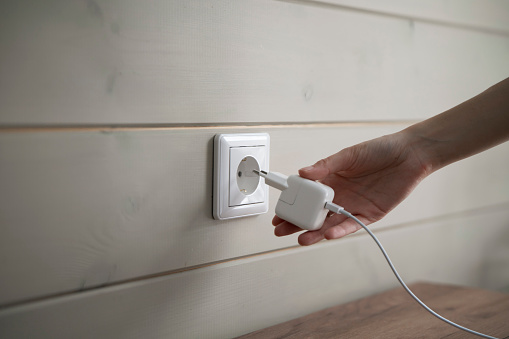 A persons hand is captured in the moment of inserting a two-pronged white electrical plug into a standard wall-mounted power socket, suggesting an indoor domestic or office setting.
