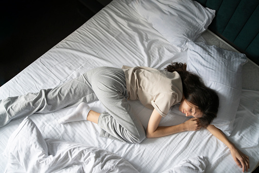 A woman is captured taking a nap, curled up in a bad wrong on a white-sheeted bed, suggesting a midday or early morning rest.
