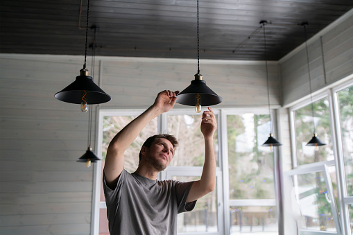 A man is captured in the act of changing a light bulb in a hanging ceiling light fixture, with natural daylight illuminating the scene.