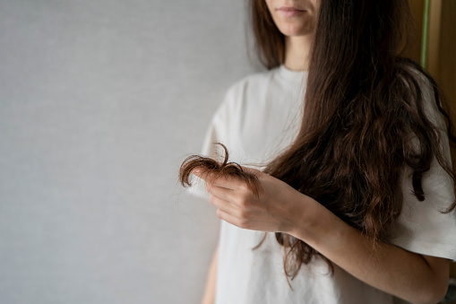 A woman appears concerned as she closely inspects the split ends of her long, wavy brunette hair, possibly considering remedies for hair damage.