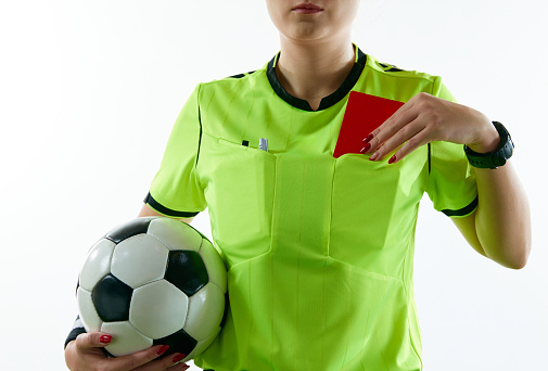Young woman, female soccer referee holding ball, stopping game and showing red card against white studio background. Concept of sport, competition, match, profession, football game, control