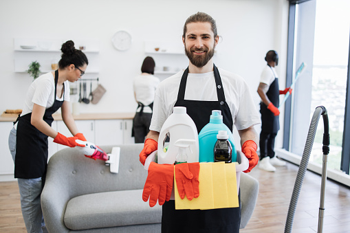 Portrait of bearded young Caucasian man professional cleaning worker holding a bucket for washing with detergents on bright kitchen studio background, copy space.