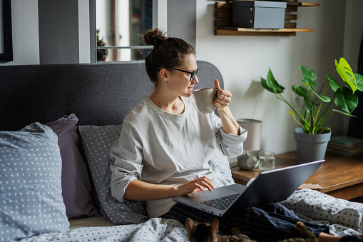 Young beautiful smiling woman drinking morning coffee and using laptop while lying in bed