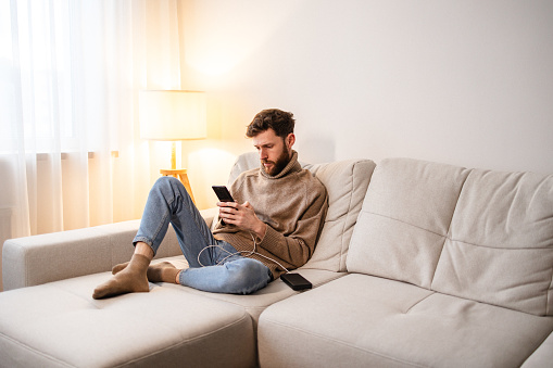 A man comfortably sitting on the sofa at home, engrossed in his smartphone. The image reflects the seamless integration of technology into everyday moments, highlighting a sense of ease and connectivity in domestic surroundings