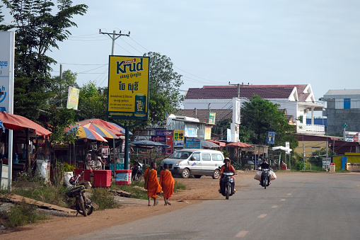 Two novice monks walking on the sideway and people riding motorcycles on road 66 in Siem Reap province, Cambodia.