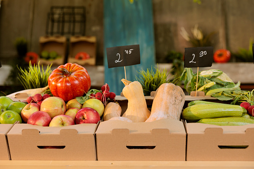 Locally grown colorful organic fruits and vegetables displayed on greenmarket stand with price tags. Close up of freshly harvested bio produce in cardboard boxes positioned on farm trade fair booth.