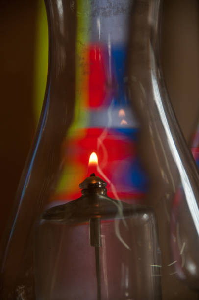 A single flame burns inside the glass globe of an oil lamp with stained glass forming a colorful background.