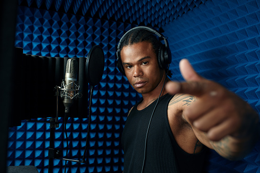 Close-up of handsome young Black singer standing in front of microphone singing song in recording studio.