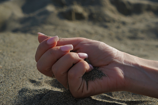 Close-up of a woman's hand releasing falling sand. Sand flows through your hand against the background of sea sand on the beach. The concept of a summer beach holiday