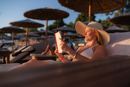 A happy woman is lying on a beach chair and reading a book on a summer day