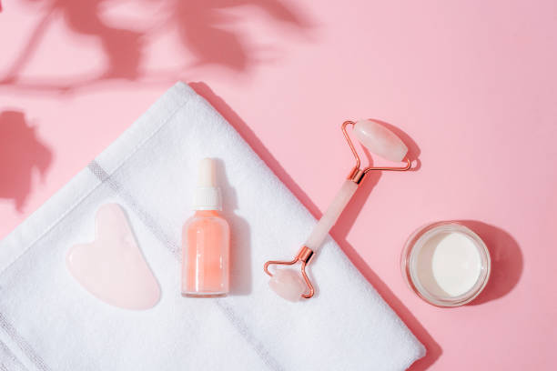istock Facial cosmetic roller, gua sha scraper, serum bottle and cream jar on a white towel on pink background with flower shadows. Home spa concept. Top view, flat lay 1939865592