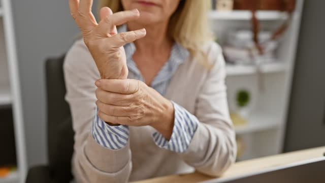 Mature blonde woman experiences wrist pain in the office, suggesting carpal tunnel syndrome or similar repetitive strain injury.