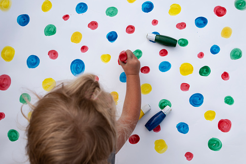 One girl paint multi coloured dots on white background. Fun play and craft of child. Multi-coloured background. Overhead view.