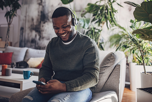 Adult man at home smiles while listening to music on headphones, engaging with his smartphone in a relaxed setting