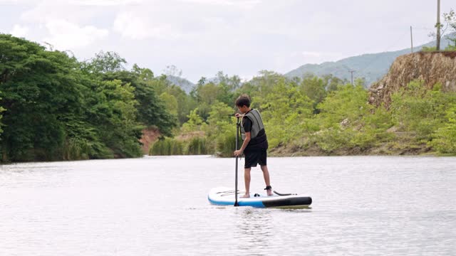 Rookie Asian athletic boy learning to play paddle board at lake. Solo outdoor water sport and travel on summer holiday.