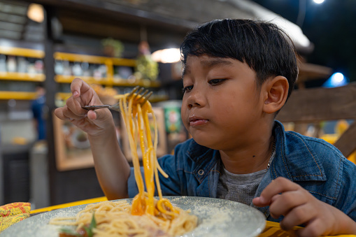 Asian little boy eat spaghetti in the restaurant on night. Pasta sauce typically made with cured pork, egg, and grated cheese.