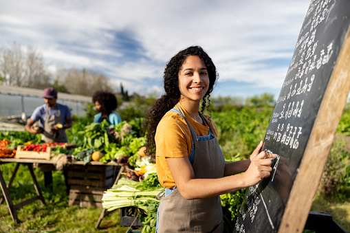 Happy Latin American woman selling her produce at a Farmer's Market and writing on a blackboard while looking at the camera smiling