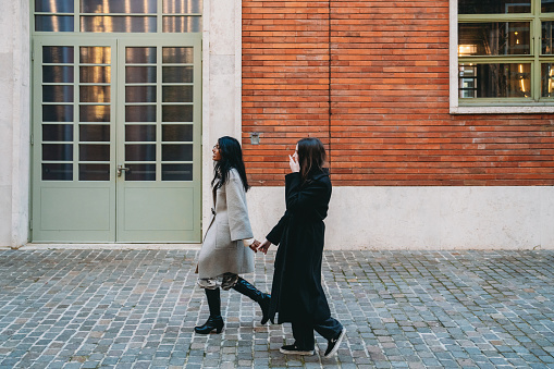 Side view of two friends walking together in the city against a brick building. They are hanging out together, having fun.