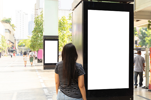 Woman standing near blank advertising lightbox on the bus stop, mock up. Blank white billboard. Banner board advertising posters. Digital advertising lightbox located in the middle of the walkway.