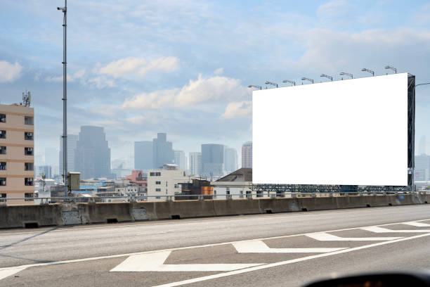 istock The billboard is rectangular and made of metal. It is painted white and has a black border. The billboard is located on the side of a busy highway. 1939989055