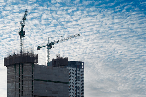 Two construction cranes are silhouetted against a blue sky with fluffy white clouds. They are lifting heavy beams of steel, which are being used to construct a new building.