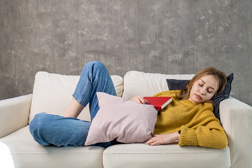 young woman in a yellow sweatshirt and blue jeans fell asleep on the couch while reading a book. reading books improves sleep