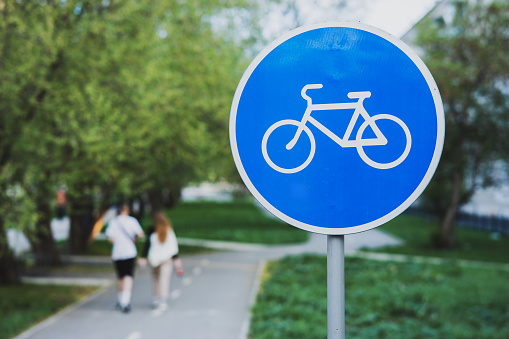a blue circular bicycle sign at the bike path. The background is blurred, people are walking