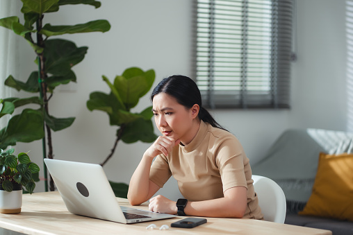 Asian woman sitting at home office, feels concerned thinking about problem with work.