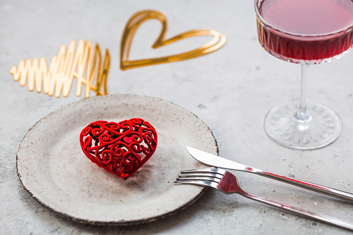 The banner. Table setting. A stylish ceramic plate, a glass of red wine, a fork and gold, red hearts on a concrete background. The concept of celebrating Valentine's Day for cafes and restaurants.