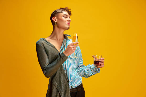 istock Overjoyed man with woman's and man's appearance holding glasses with alcohol drinks against vivid yellow background. 1940022039