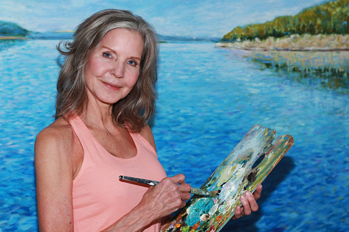 Pretty Older Fit Toned shoulder length gray hair woman wearing a peach sleeveless top standing in front of impressionistic lake landscape painting holding a paintbrush and palette smiling at camera.