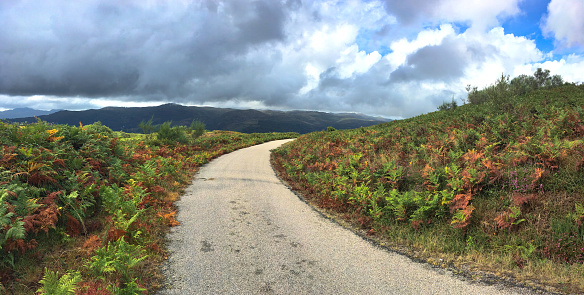 Panoramic Horizontal photo of a Tarmac Road in the Middle of the Mountains Towards Nowhere, with Heavy, Dark Clouds of Rain