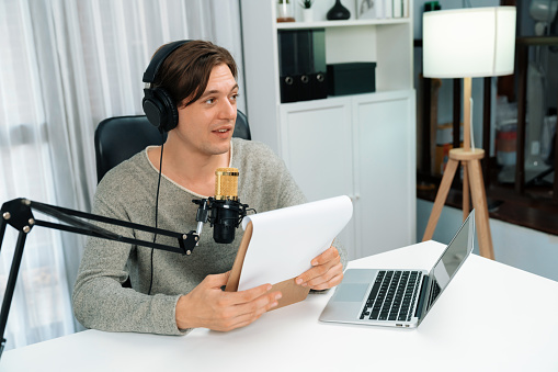 Host channel in smart broadcaster with talking show on live social media streaming with script note reading to listeners, wearing headphones to record video streamer at modern home studio. Pecuniary.