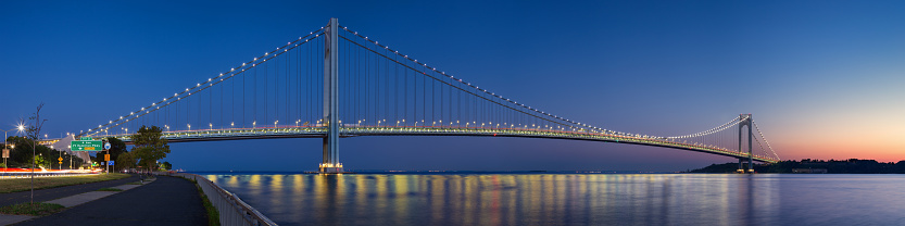 High resolution stitched panorama of the Verrazano-Narrows Bridge at Red Orange Yellow Blue Sunset. The bridge connects boroughs of Brooklyn and Staten Island in New York City. It was built in 1964 and is the largest suspension bridge in the USA. Historic Fort Wadsworth is next to the bridge foot. The photo was taken from the Shore promenade in Bay Ridge Brooklyn. Canon EOS 6D full frame censor camera. Canon EF 50mm F/1.8 II prime lens. 4:1 Image Aspect Ratio. This image was downsized to 50MP. Original image resolution is 65MP or 16146 x 4036 px.