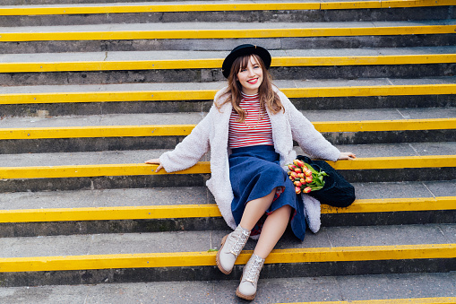 Portrait of young smiling fashion woman with fresh tulips in her net bag sitting on stairs at the city street. Confident female. Urban street fashion. Springtime mood. Selective focus. Copy space.