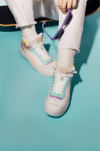 Close up female legs in white jeans and retro style high-top multicolor sport sneakers shoes sitting on the skateboard on turquoise blue background. Vintage retro fashion style of 80s - 90s vibes.