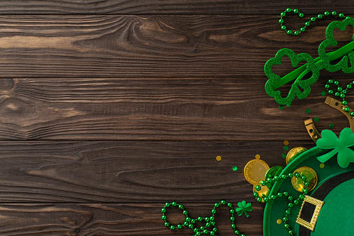 Leprechaun's Legacy: top view St. Patrick's Day essentials â leprechaun's hat, themed glasses, lucky horseshoe, gold coins, trefoils, confetti, beads on wooden surface. Set mood for joyous celebration