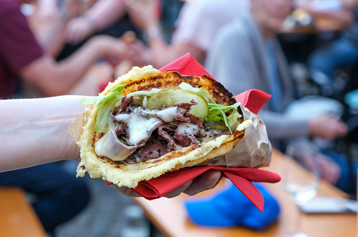 Close-up of a delicious pastrami sandwich with sauce and vegetables in a potato waffle held by a woman at an outdoor festival, concert, or street food festival, selective focus