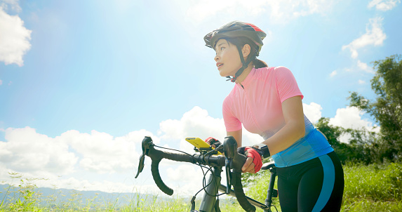 enjoy sport or healthy lifestyle concept - asian young woman wearing helmet is setting tracker app through smart phone and standing by a bicycle on the road