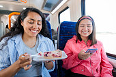Two young latin friends women enjoying traveling in public train transportation, using phone and eating pie