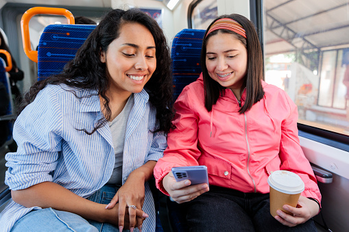 Two young latin female students or commuters traveling on public train transportation in the city and using mobile phone. Technology and fun on the go