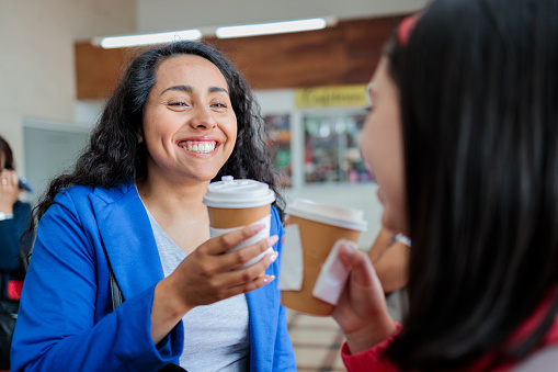 Two young smiling latin women commuters waiting for public train transportation in the station while drinking hot coffee