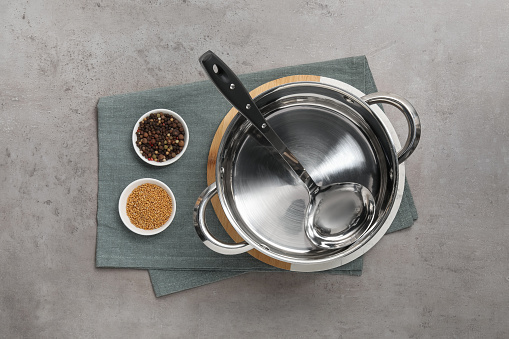 Steel pot with ladle and spices on grey textured table, top view