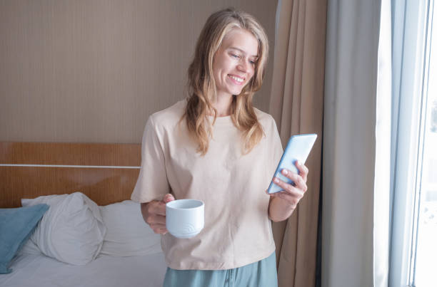 istock Woman standing beside the window using mobile phone holding coffee cup 1940383909