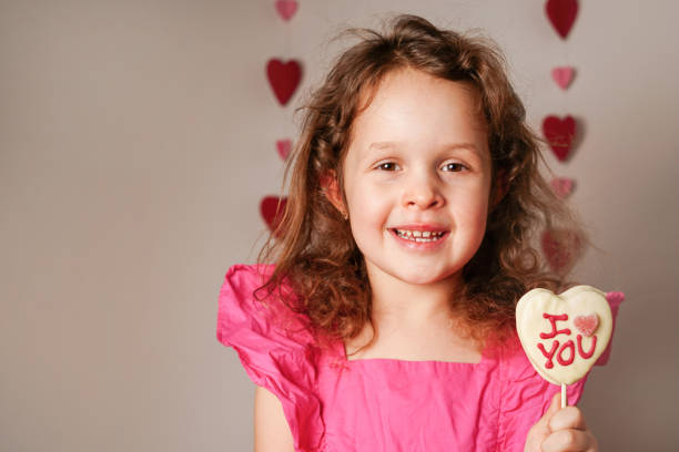 menina da criança no vestido cor-de-rosa segurando o coração do dia dos namorados em forma de doce com chocolate branco. conceito de amor - child valentines day candy eating - fotografias e filmes do acervo