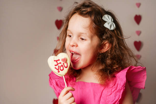 ピンクのドレスを着た女の子が、バレンタインデーのハート型のキャンディーをホワイトチョコレートで食べています。愛のコンセプト - child valentines day candy eating ストックフォトと画像