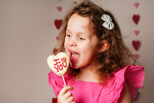 Child girl in pink dress eating Valentine's day heart shape candy with white chocolate. Love Concept