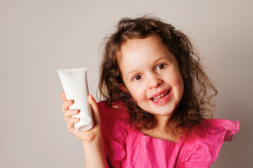 Child little child girl holding white cosmetic cream tube and smiling. Daily children beauty product