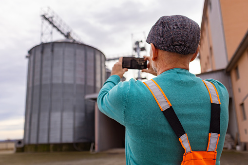 With a mobile phone in hand, a worker in overalls documents a corn silo scene.