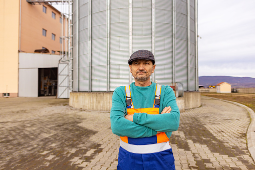 A worker in overalls stands proudly before a towering silo, embodying strength and dedication in an industrial setting.